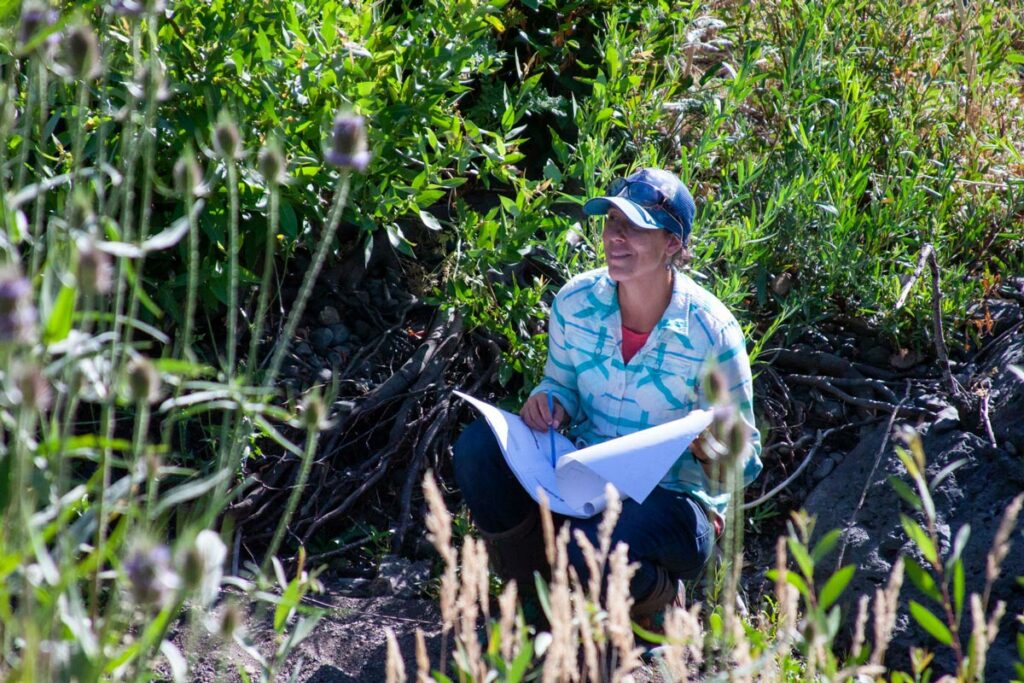 Tuli Potts, SNC's new Area Representative for the North Subregion, crouches in dry streambed with green bushes behind her. She is wearing a blue baseball cap a plaid shirt, jeans, and boots and is holding a pen in her hand that is resting on a large sheaf of papers that is balanced on her legs.