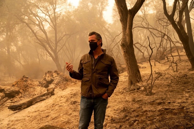 Governor Newsom stands wearing a mask with burned trees and charred ground behind him that is made hazy by smoke.