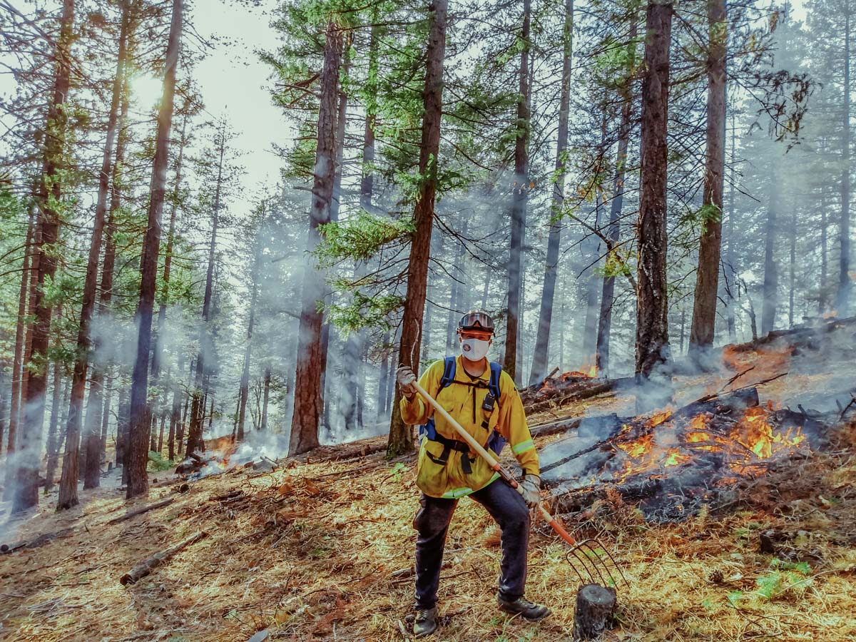 man in a hard had, mask, and shovel poses on front of small flames burning behind him in a forest