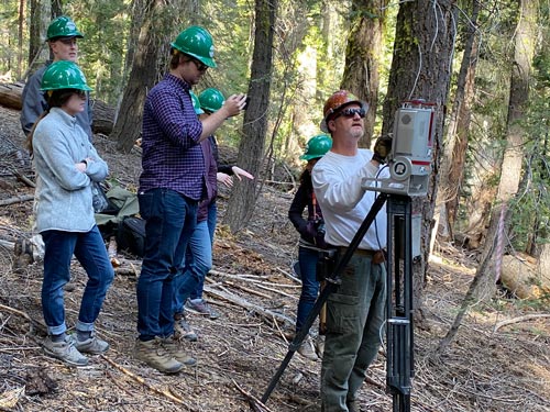 A man standing in a forest wearing a hard hat is looking into a small machine on top of a tripod with a few other people in hard hats standing by