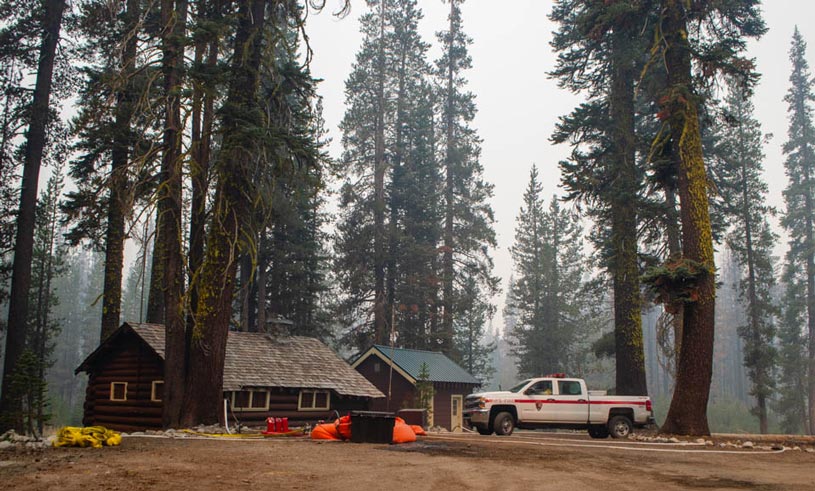 Fire truck parked next to two cabins in a smoky forest. Firefighting equipment is staged next to the cabins.