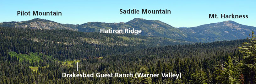 Forested landscape with three peaks in the background: Pilot Mountain, Saddle Mountain, and Mount Harkness. Flatiron Ridge is in the middle ground, and Drakesbad Guest Ranch (Warner Valley) is near the foreground.