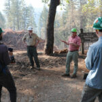 group of people wearing hard hats stand in circle in a forest listening to a man talk