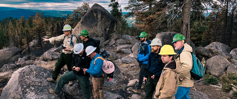8 people wearing hard hats stand atop a rocky mountain ridge, talking and looking out across a landscape