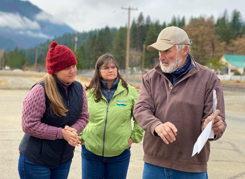 Three people standing outside on asphalt, man on the right holds out a piece of paper while talking, the other two women are leaning in and listening. Forested hillside with telephone wires is behind them and a mountain is in the background