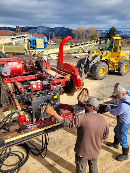 two men stand outside in front of a saw machinery with a tractor and mountains in the background