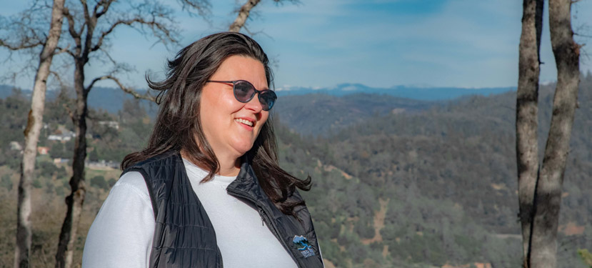 woman with long dark brown hair, wearing a black vest and white long-sleeved shirt and sunglasses is looking out toward the right with a small open smile. Hillside with a few houses and a small mountain is in the background.