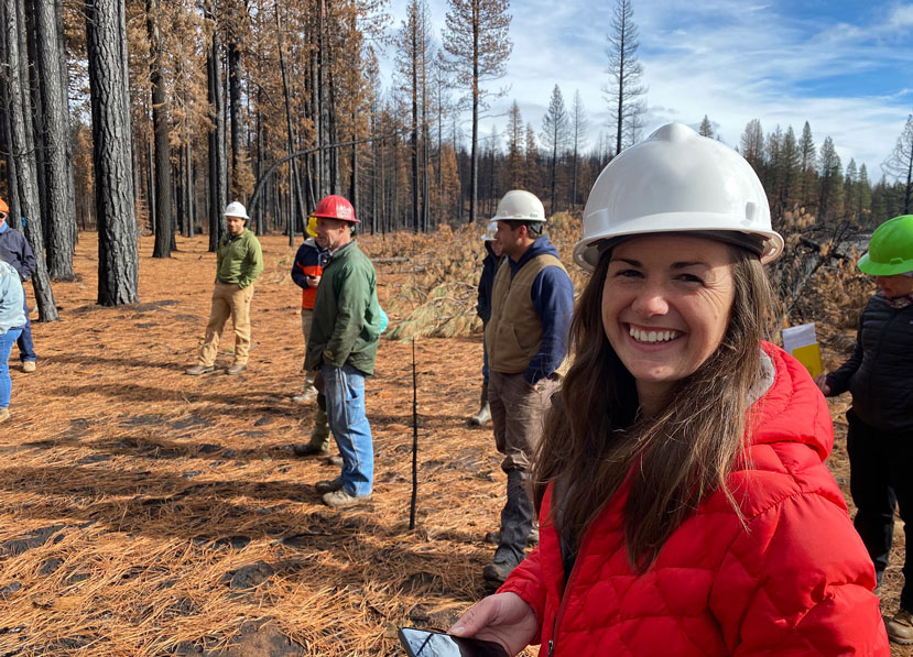 woman wearing a hard hat, smiling at the camera in the foreground, behind her other people stand in hard hats with burned trees in the background and burned pine needles on the ground