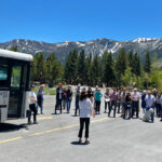group of people stand in the road next to a bus that says Mammoth with trees and mountains in the background