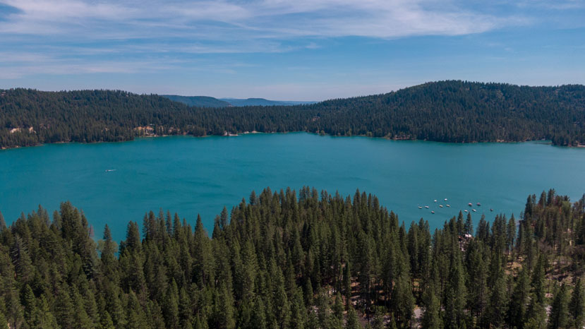low aerial looking out across a teal blue reservoir with green pine trees in the foreground and background.