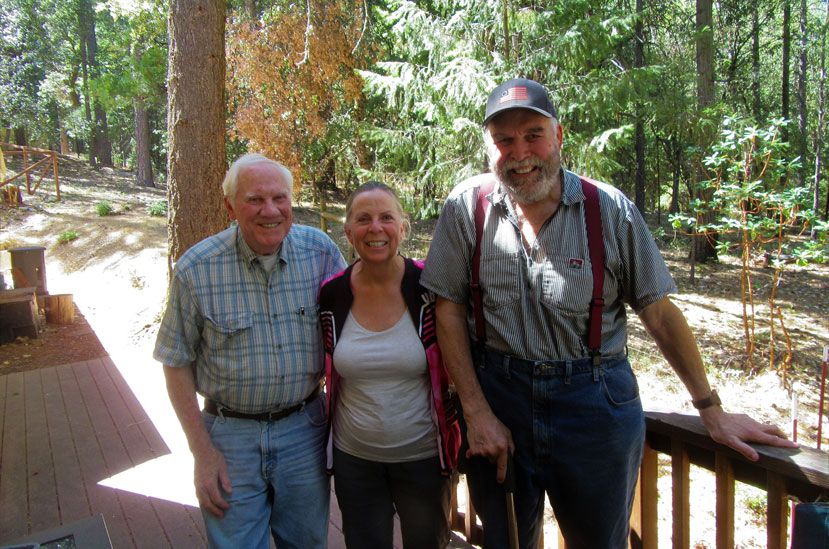 Three people (an older man, a woman, and another man) stand on a porch smiling facing the camera with a forest behind them