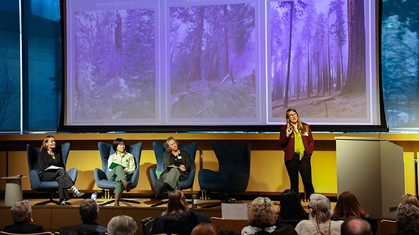 a woman standing on a stage in front of a screen with photos of trees, three people sitting on the stage to the side