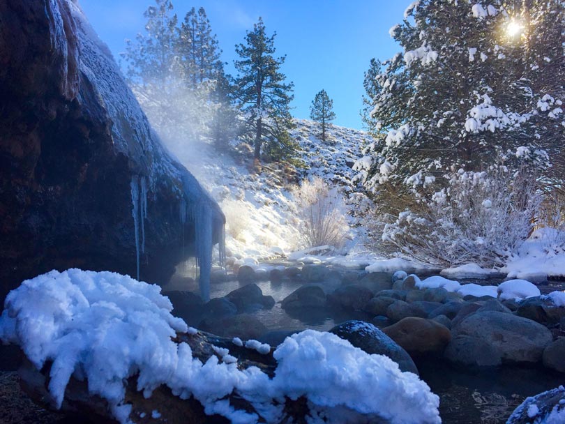 snow covers rocks and trees surrounding a small body of water with steam rising from it