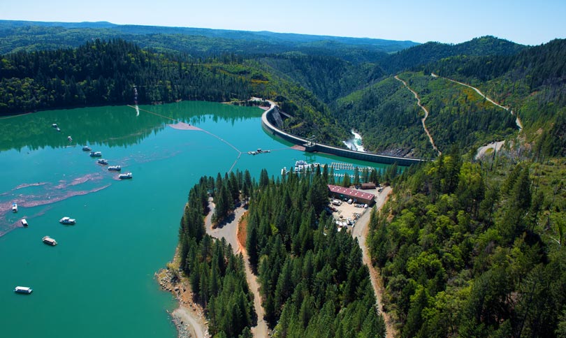 Aerial of a dammed lake with teal, blue-green water with a few boats. It's surrounded by vibrant green pine trees that are bisected by a few roads.