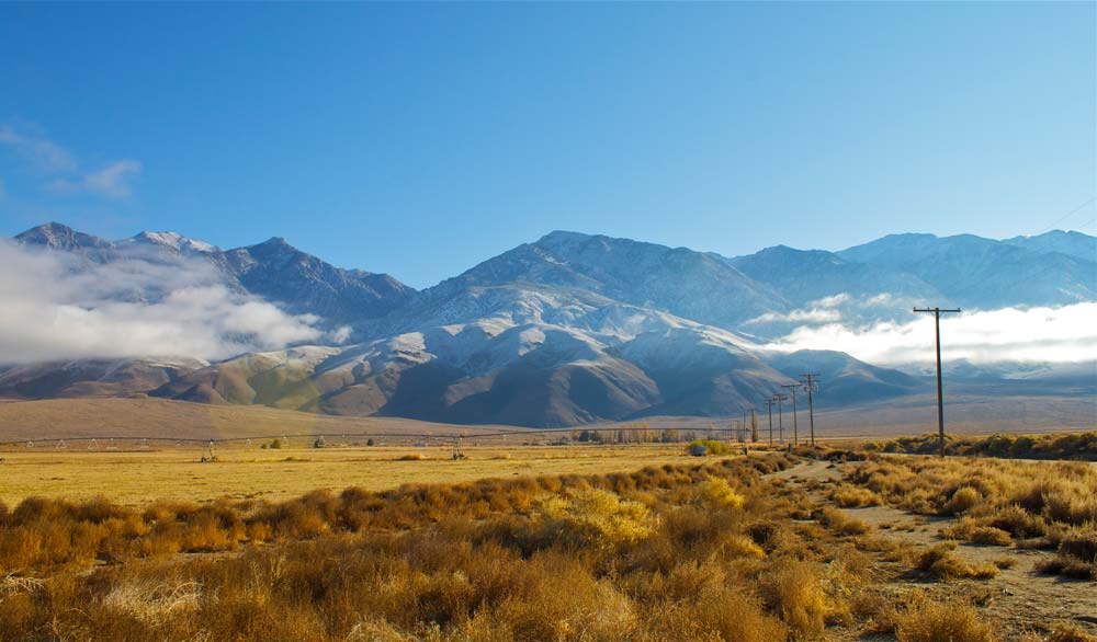 golden brown shrubbery and grass extend into the background toward large, craggy, snowy mountains. Telephone poles on the right side of the image fade into the mountains in the background