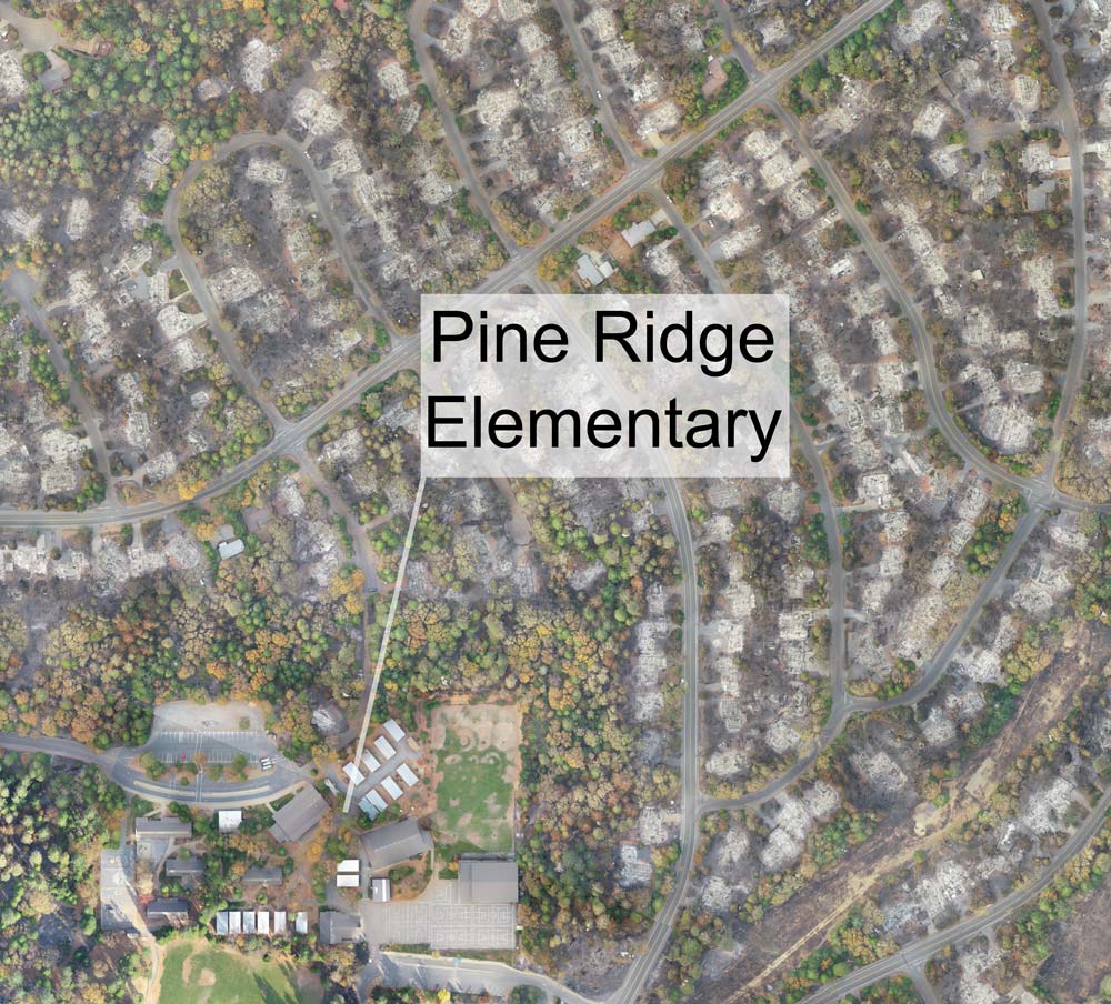aerial showing neighborhood mostly burned, but Pine Ridge School is still standing with green trees in the bottom left of the frame