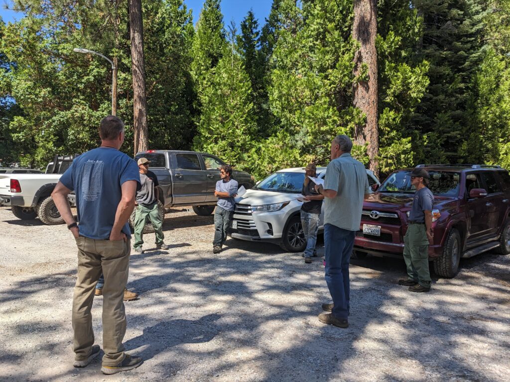 Seven people are gathered in a circle, discussing the project in a gravel parking lot.