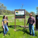 Two people standing in front of a preserve sign with green grass and oak trees in the background.