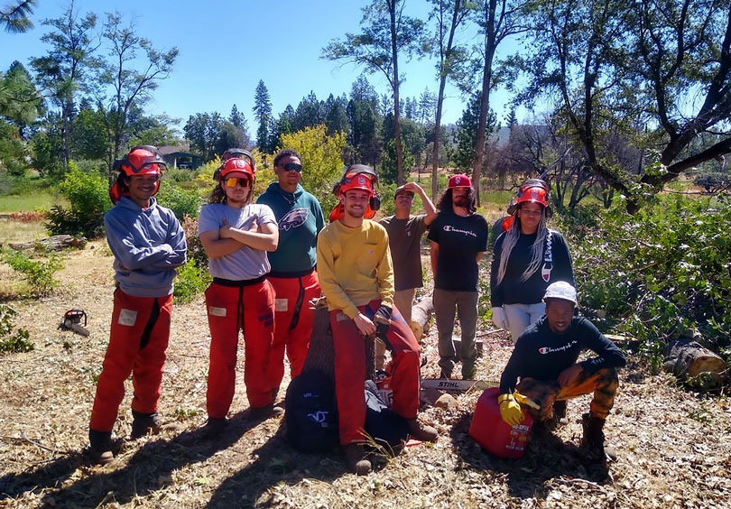 8 young adults pose, standing on dirt surrounded by trees. Many are wearing orange long pants and hard hats with face shields.