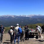 Twelve people gathered around an overlook with a picnic table. Conifer forests and mountains are in the background.