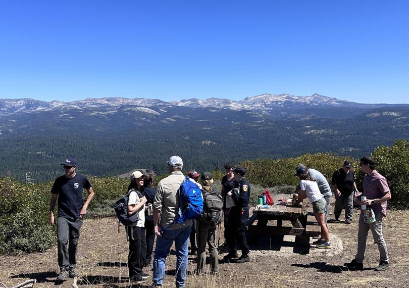 Twelve people gathered around an overlook with a picnic table. Conifer forests and mountains are in the background.