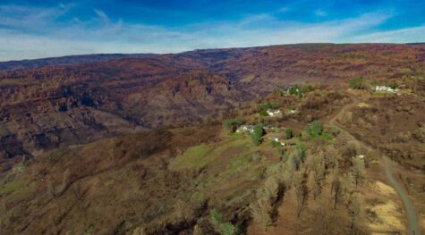 Aerial looking out across a vast burned hillside covered in mostly brown and black with a handful of white structures and green trees still remaining in one area.