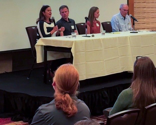 A panel of four people seated at a table speak into microphones to an audience.